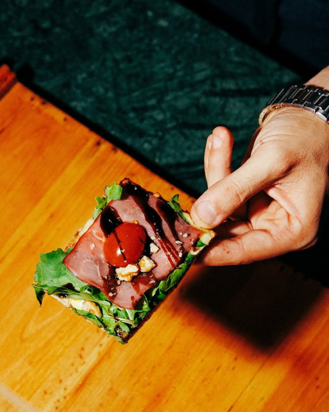 Hand holding a Roman-style pizza slice with prosciutto, rucola and balsamic glaze